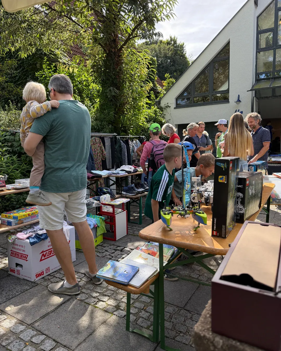 Besucher an Biertischen mit Kleidung, Spielen und Lego-Technic-Kartons vor einem Haus