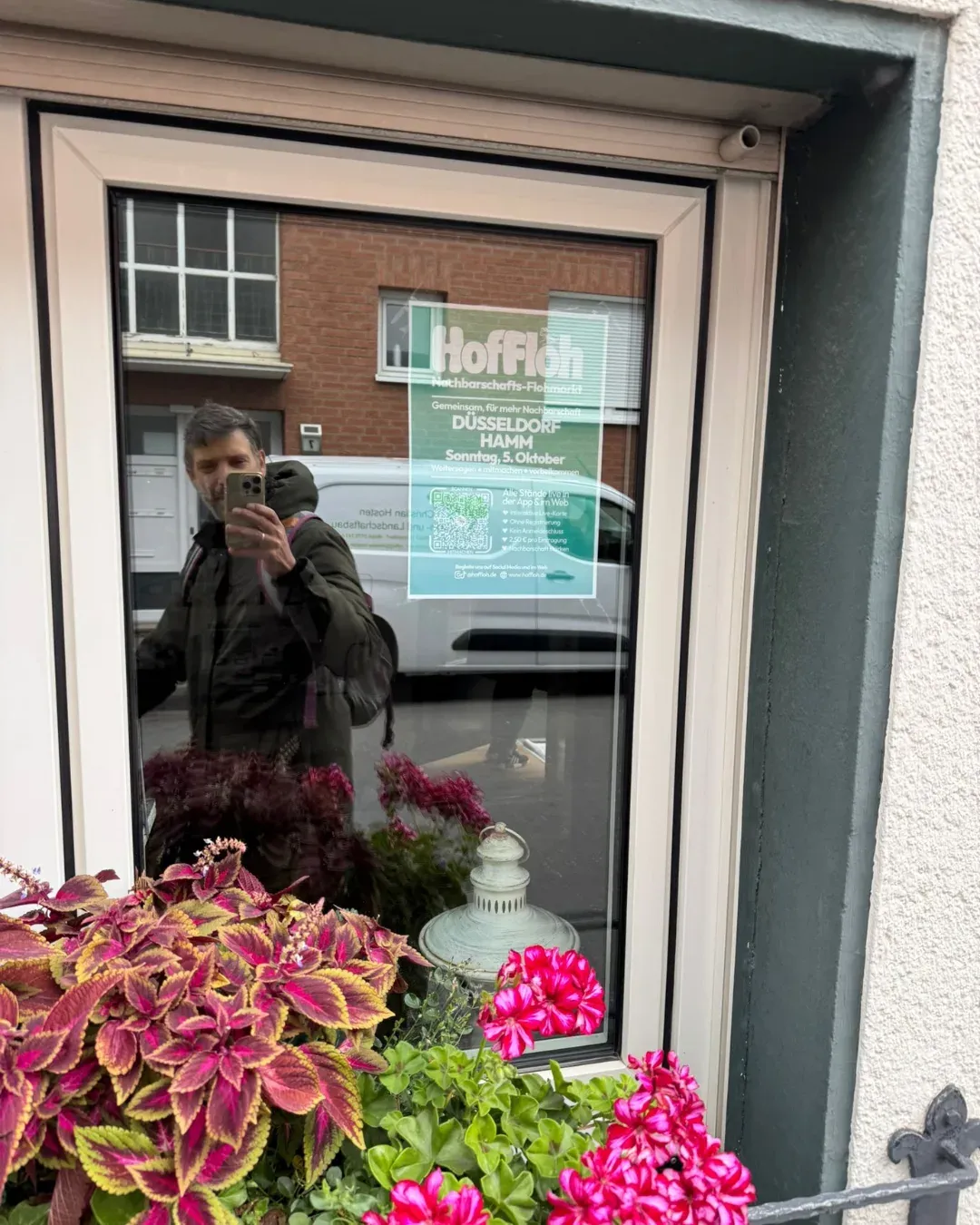 HofFloh-Plakat für Düsseldorf-Hamm in einem Fenster, davor Blumen und eine Person, die sich spiegelt und fotografiert