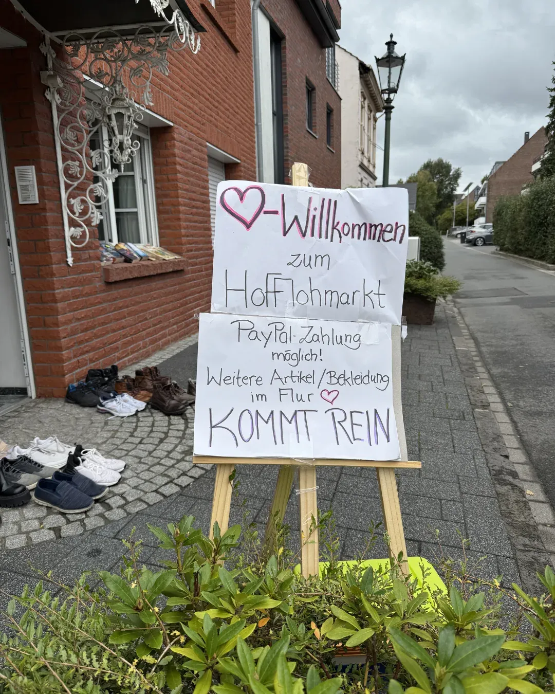 Schild „Willkommen zum Hofflohmarkt“ mit Hinweis auf PayPal-Zahlung, daneben Schuhe vor einem Haus