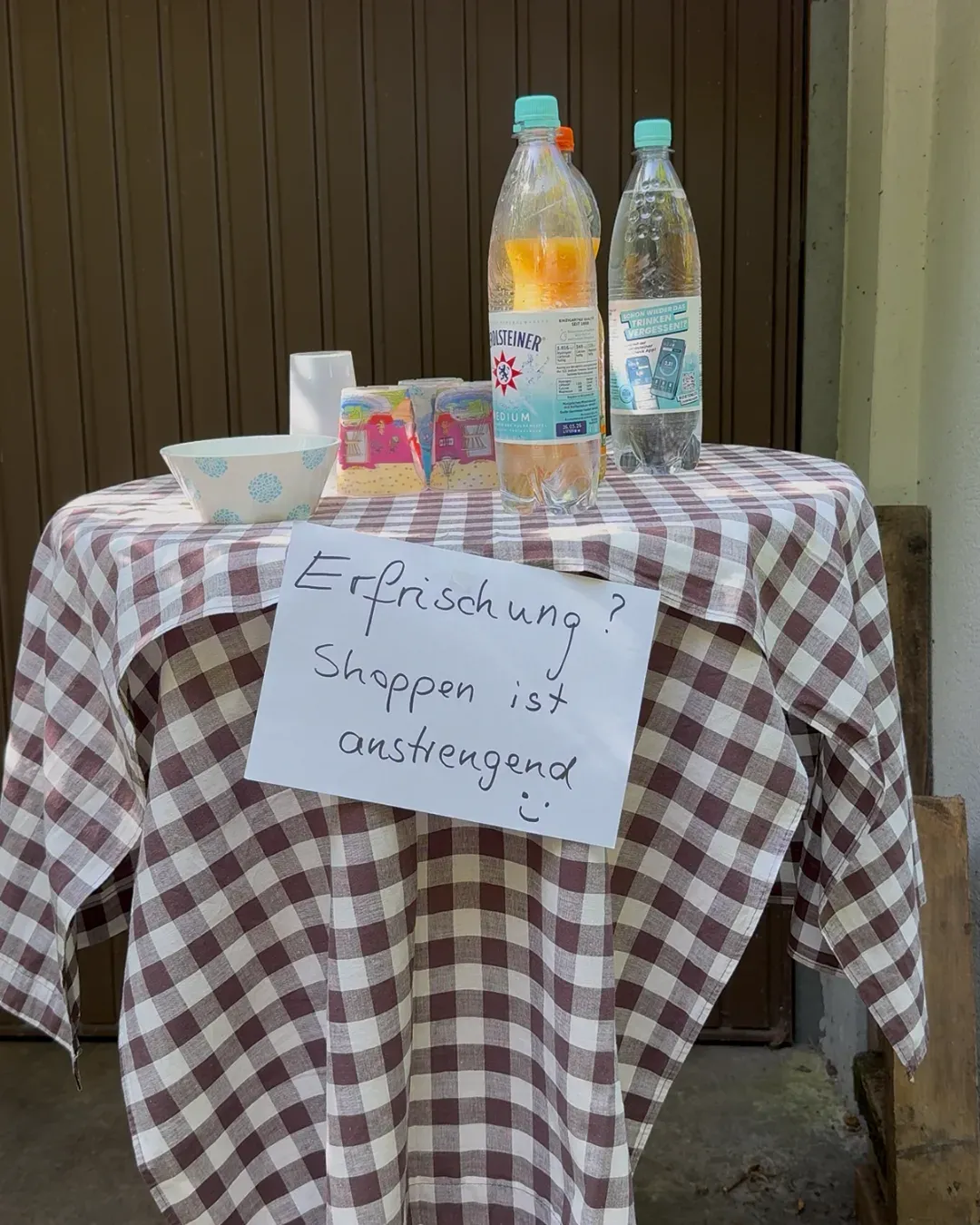 Tisch mit kariertem Tuch, Wasserflaschen und Bechern, Schild „Erfrischung? Shoppen ist anstrengend“