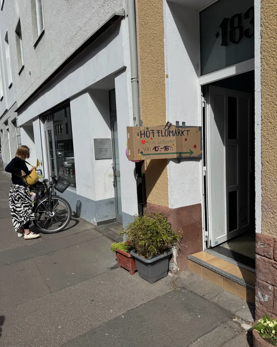 Pappschild „Hofflohmarkt“ an einer Hauswand, daneben eine Frau mit Fahrrad auf dem Gehweg