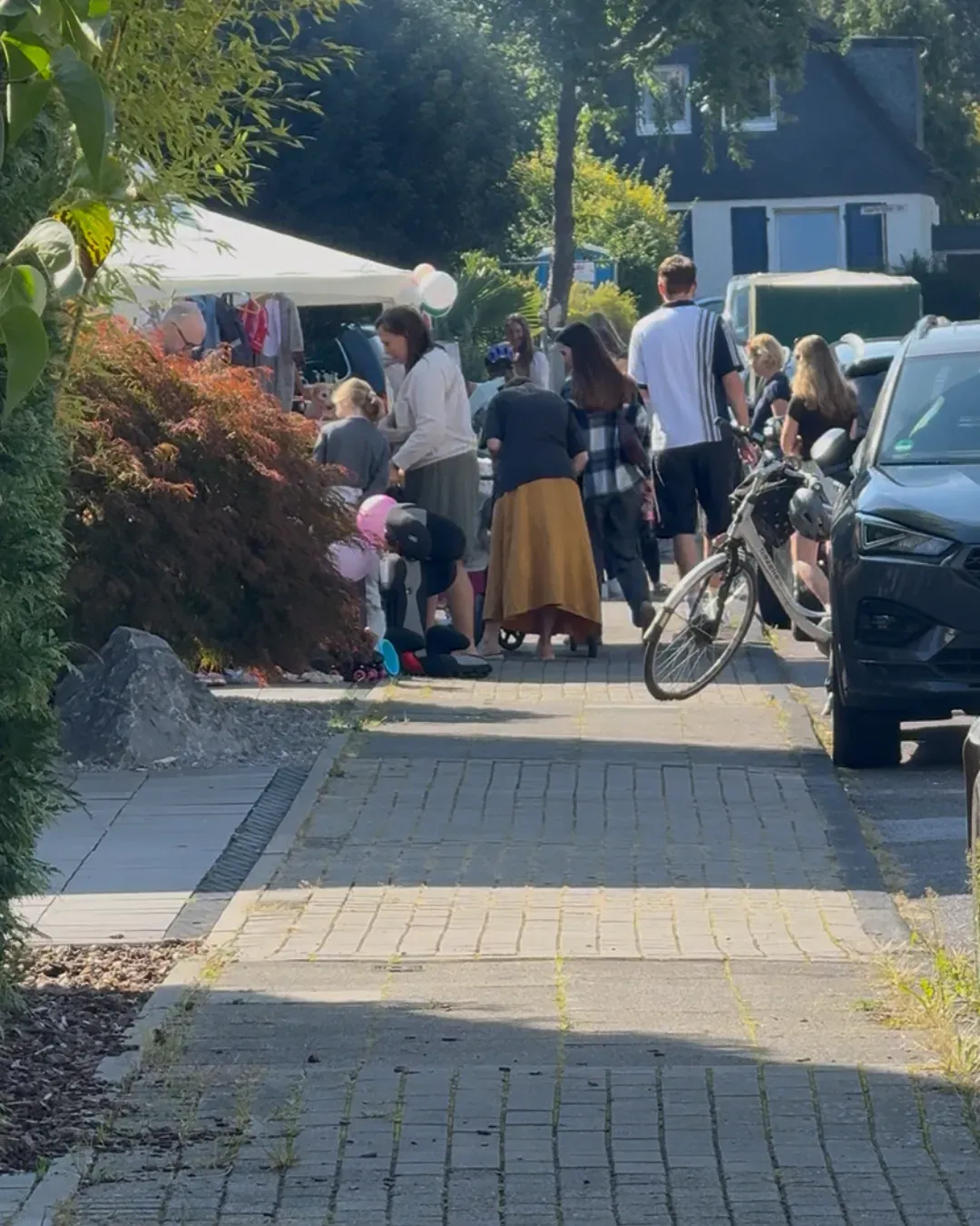 Menschen stöbern an Ständen auf einem Gehweg, daneben ein Fahrrad und parkende Autos