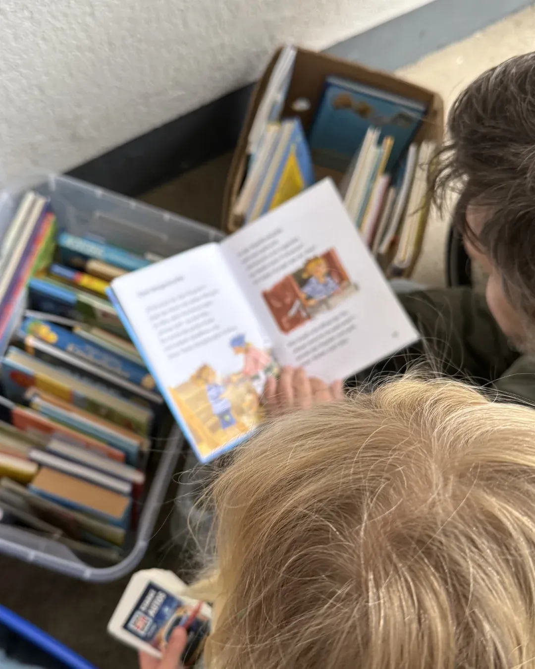 Kind und erwachsene Person blättern in einem Kinderbuch neben Kisten voller Bücher