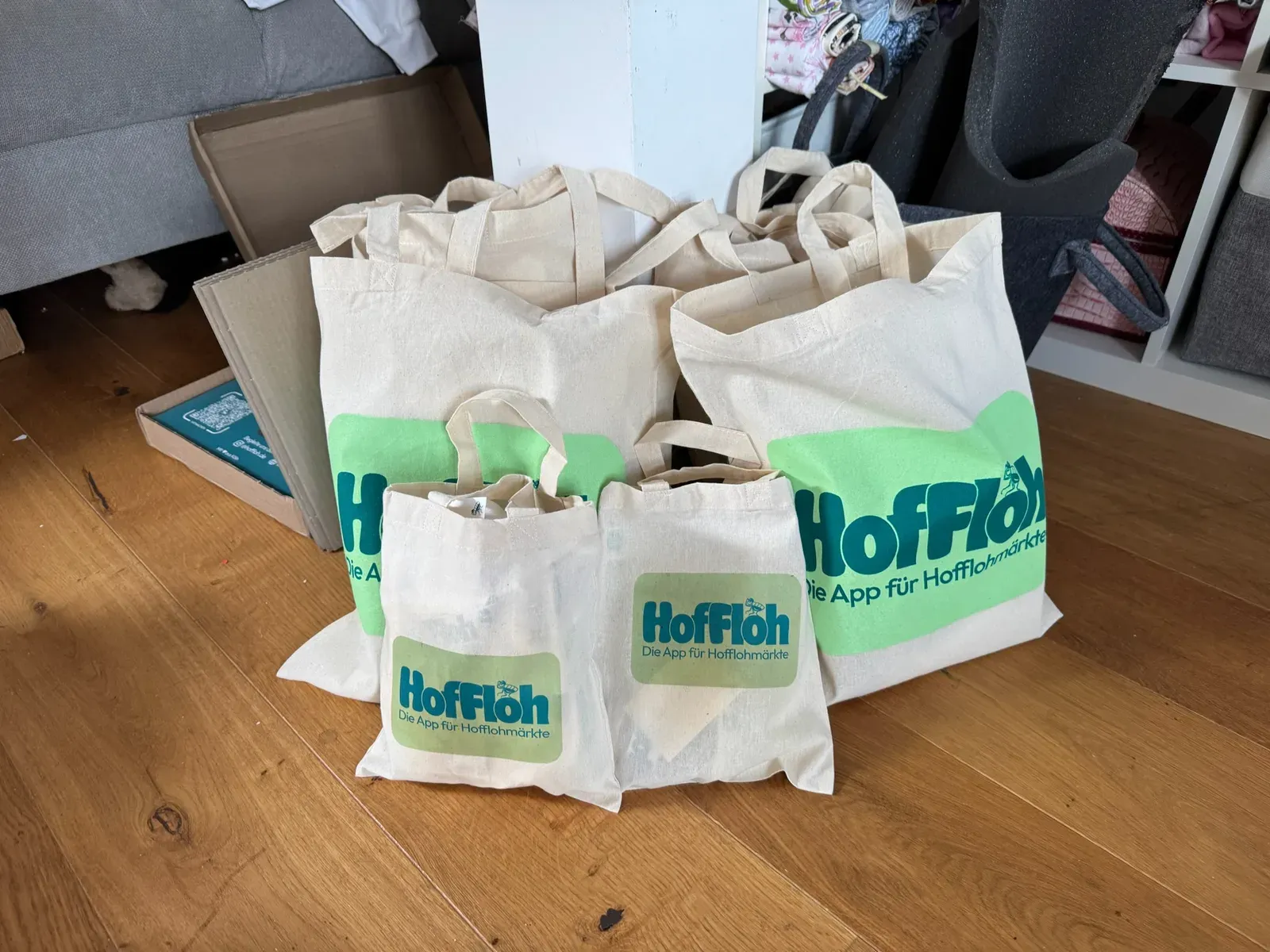 Vier Stoffbeutel mit HofFloh-Logo und „Die App für Hofflohmärkte“ auf einem Holzboden