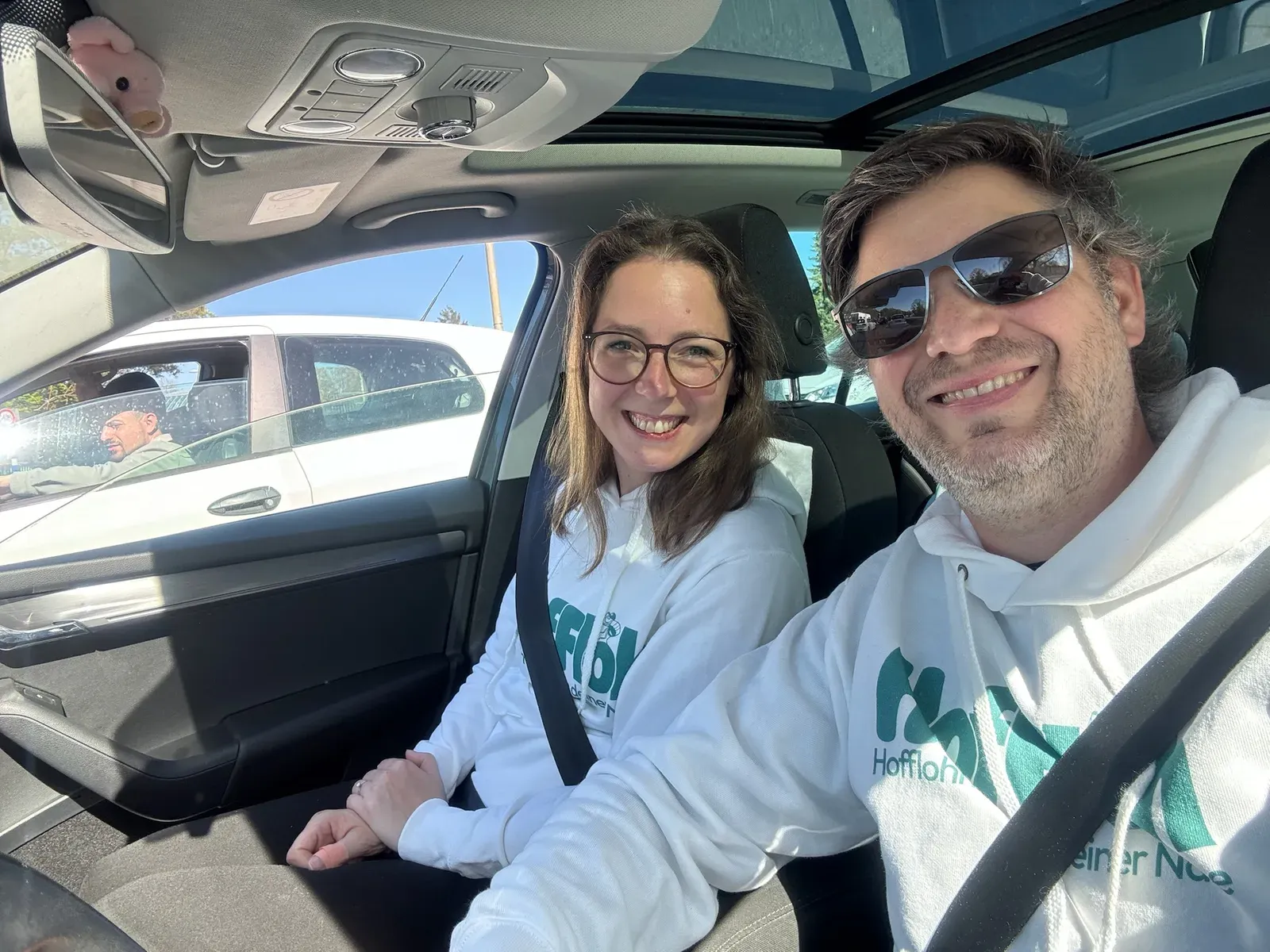 Andrea und Basti in weißen HofFloh-Hoodies lächelnd im Auto