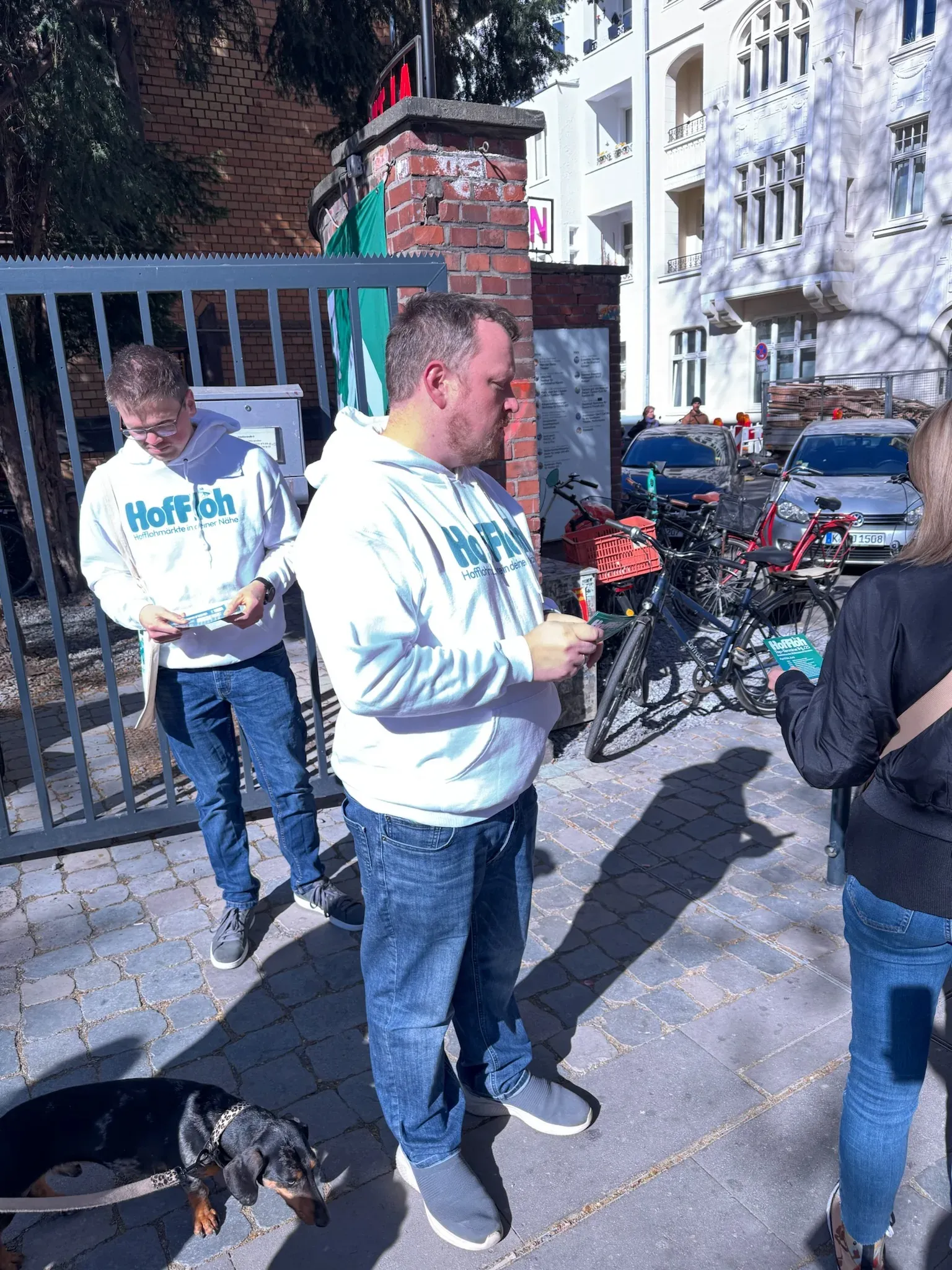 Zwei Männer in HofFloh-Hoodies verteilen Flyer vor einem Tor mit Backsteinpfeiler