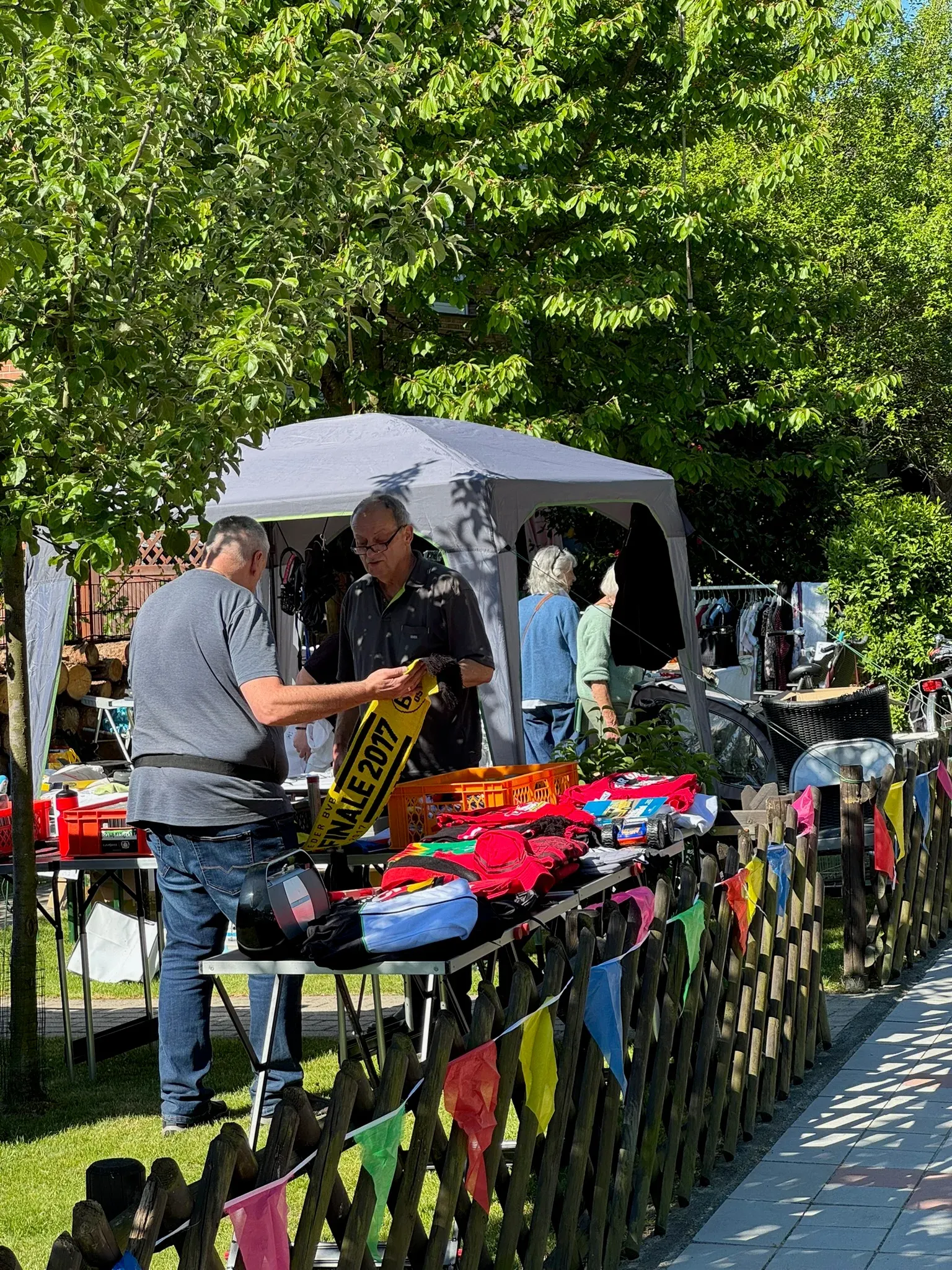 Zwei Männer an einem Stand mit Pavillon, einer hält einen gelben Fanschal, am Zaun bunte Wimpel