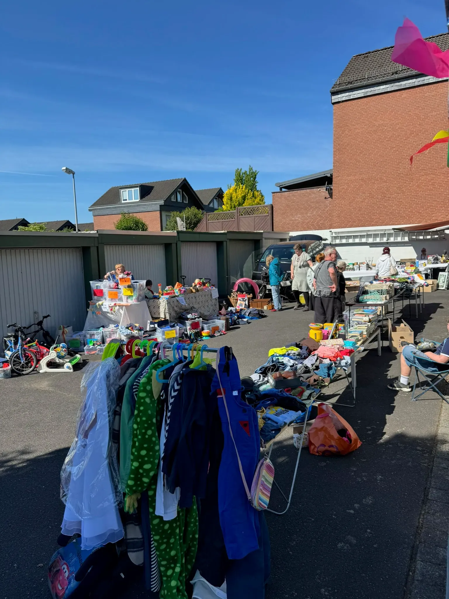 Garagenhof voller Stände mit Kinderkleidung am Ständer, Büchern und Spielzeug unter blauem Himmel