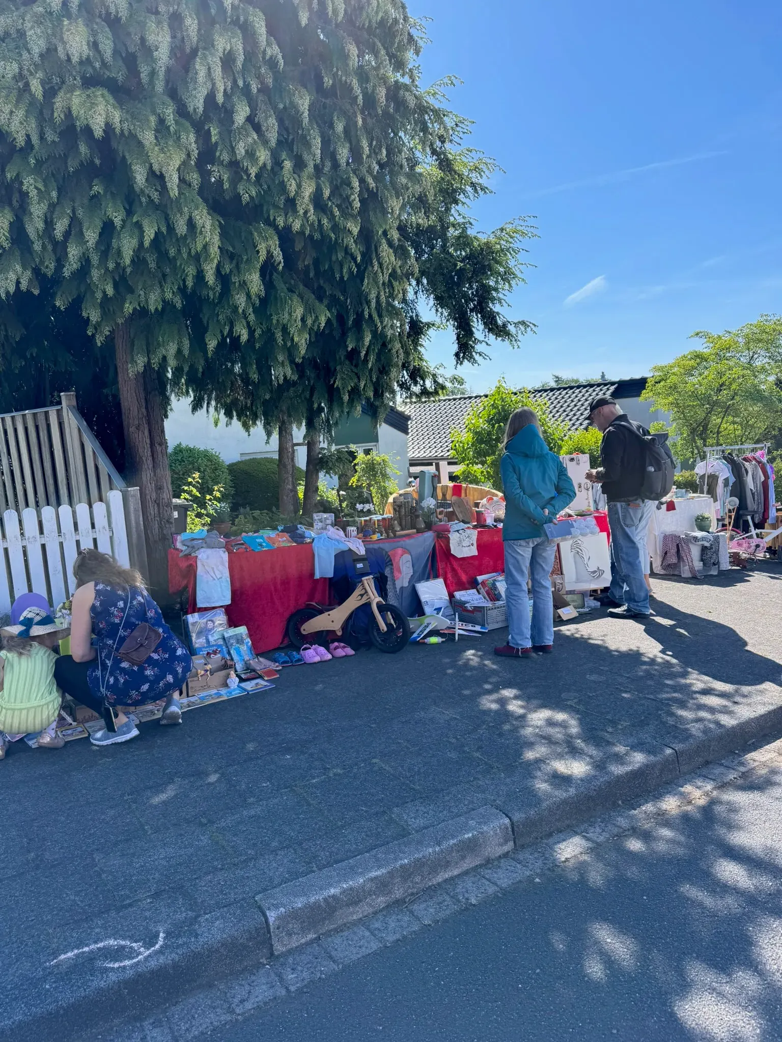 Besucher:innen stöbern an einem Stand mit roten Tischdecken, Laufrad und Kindersachen unter einem Baum