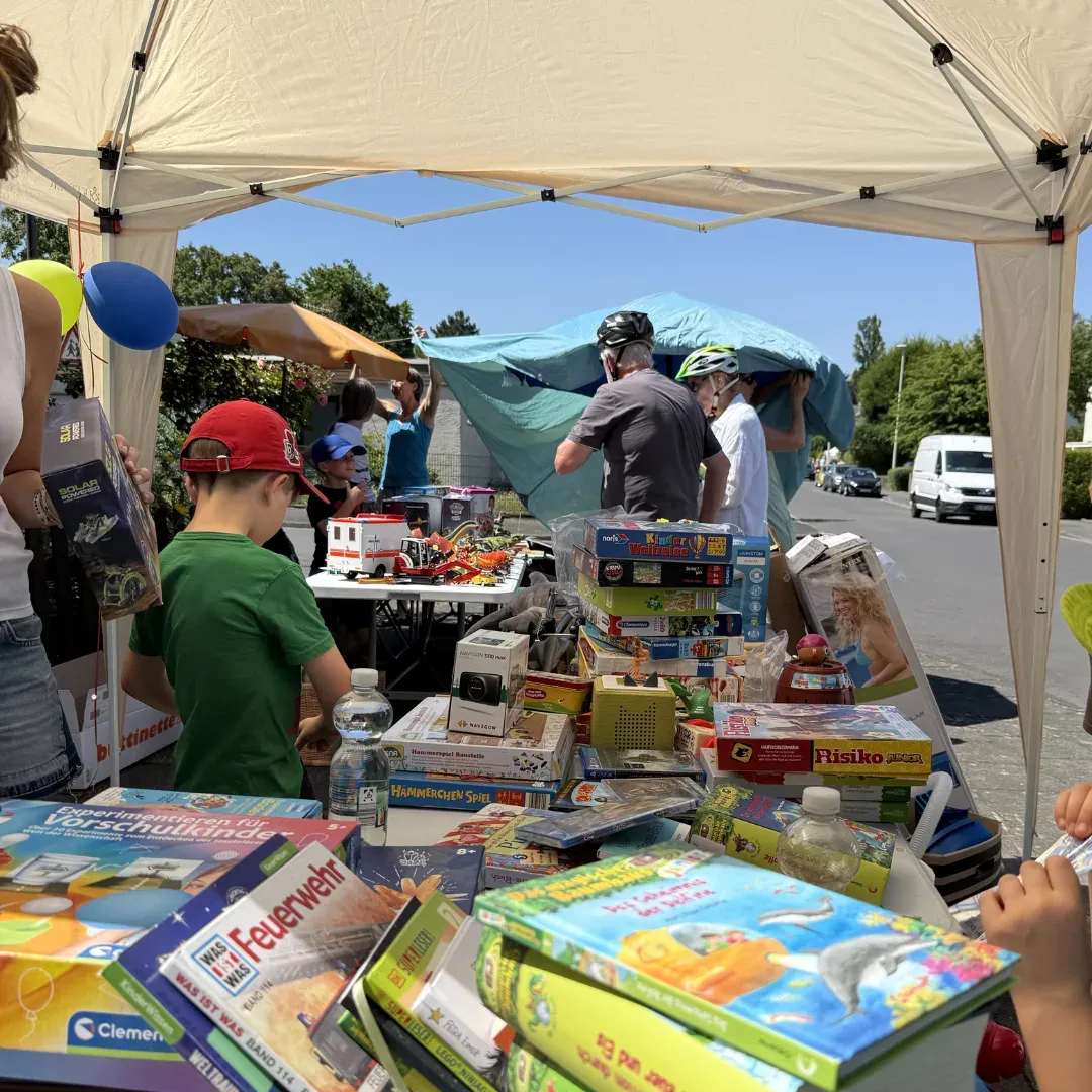 Pavillon-Stand mit Spielen und Kinderbüchern, dahinter Besuchende mit Fahrradhelmen an einer sonnigen Straße