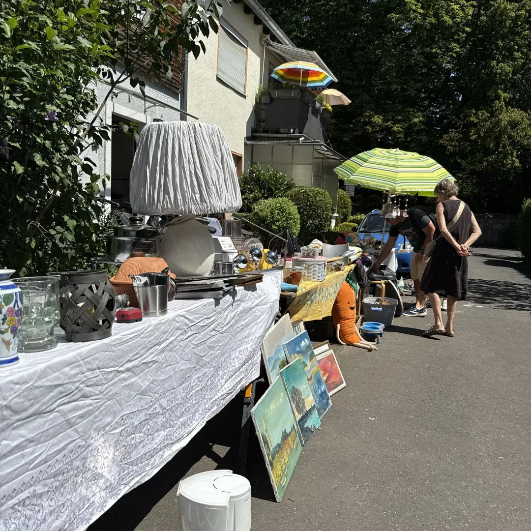 Stand mit Lampe, Geschirr und Gemälden in einer Einfahrt, zwei Besuchende schauen unter einem Sonnenschirm