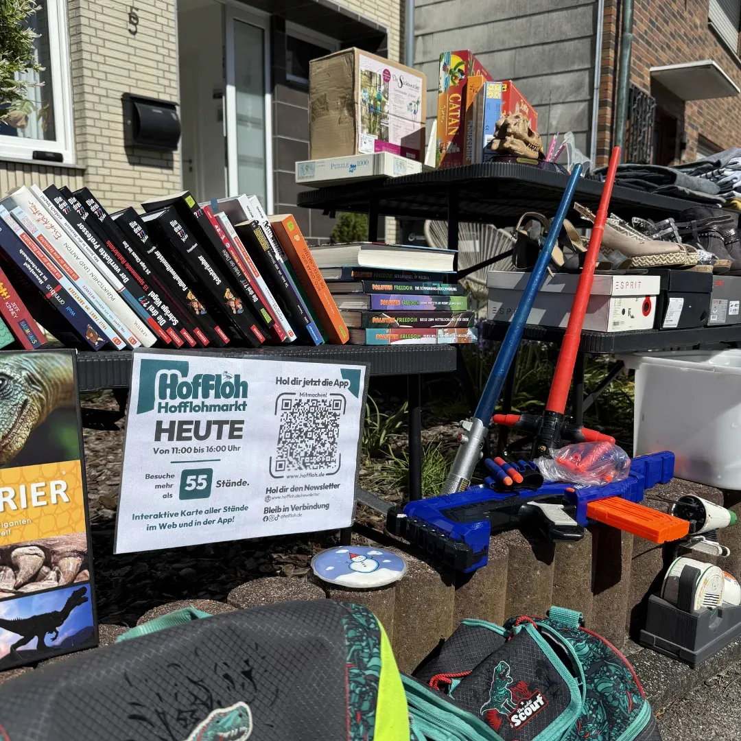 Stand mit Büchern, Spielen und Spielzeug vor einem Haus, davor ein HofFloh-Schild „Hofflohmarkt heute“