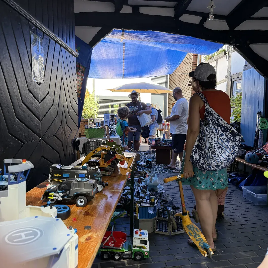 Überdachte Hofeinfahrt mit Spielzeug auf einem Biertisch, Besuchende stöbern unter einer blauen Plane