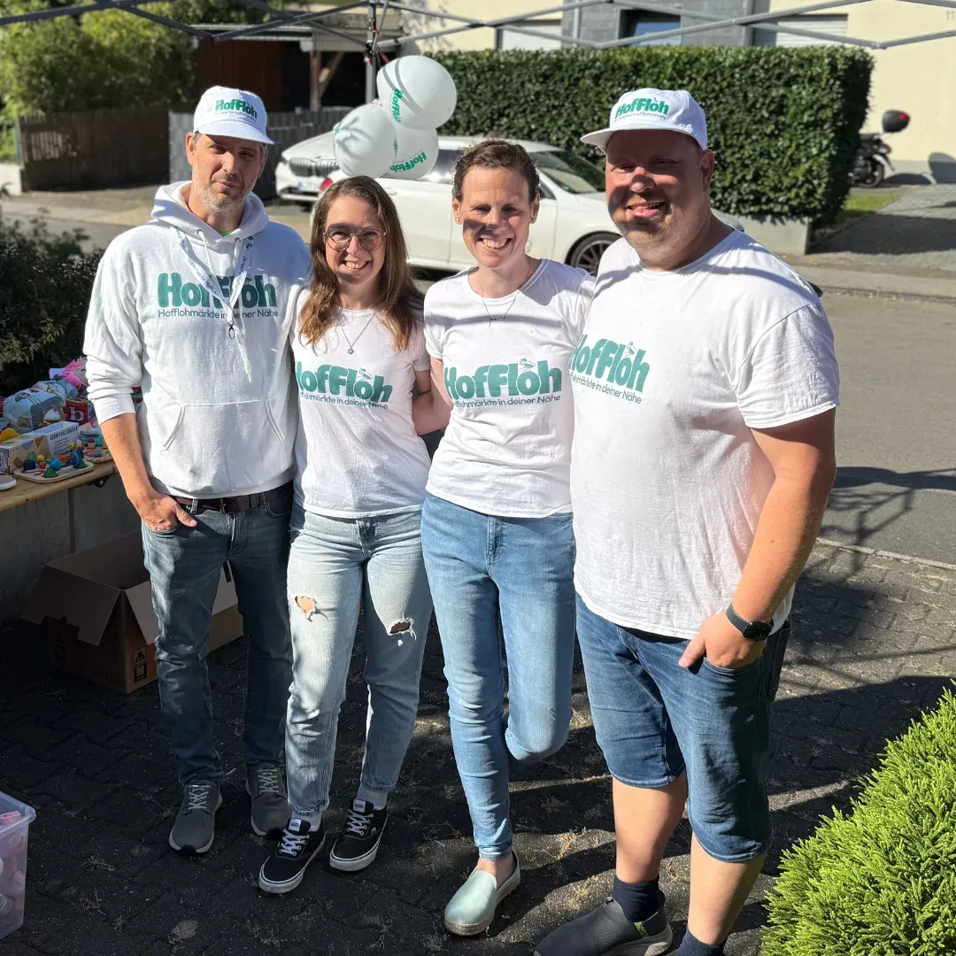 Vier Personen in HofFloh-Shirts und -Kappen stehen lächelnd in einer Einfahrt neben einem Stand