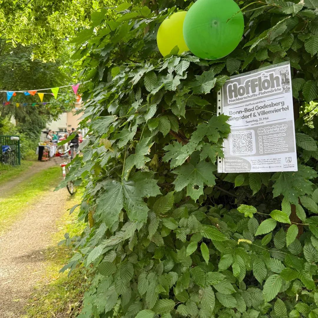Grüne Luftballons und HofFloh-Plakat an einer Hecke, im Hintergrund ein Stand mit Wimpeln